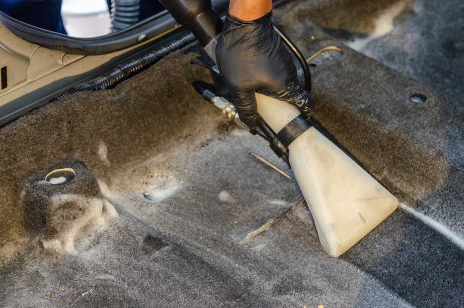 A person wearing a black glove using a handheld steam cleaning tool to deep clean the carpeted floor of a vehicle's interior, focusing on removing dirt and stains. The scene shows the nozzle of the st